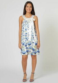 Vestido floral con flores azules sobre un fondo blanco, escote de satén y detalle de lazo. Corte en A, a la altura de la rodilla, combinado con sandalias azules.