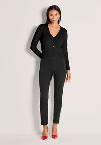 Schwarze, gerippte Strickjacke mit V-Ausschnitt und Knöpfen, kombiniert mit schwarzen, schmal geschnittenen Hosen und roten spitzen Absatzschuhen.