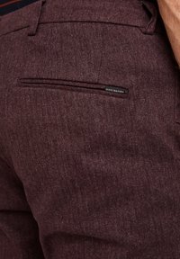 Bordowe spodnie z wzorem jodełki, z tylną kieszenią i małą markową metką. Faktura wydaje się tkana i gładka.