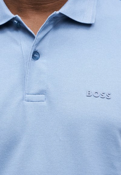 Polo bleu clair texturé avec deux boutons et logo "BOSS" sur la poitrine droite, porté par une personne avec le cou et la clavicule visibles.