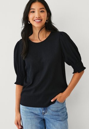 Jonge vrouw die lacht, met een zwarte, gestructureerde blouse met opgeblazen, geplooide mouwen en lichtblauwe spijkerbroek, hand in de zak.