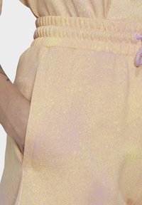 Ruka uvnitř kapsy světle žlutých tepláků s elastickým pasem a šňůrkou, která ukazuje část tetované předloktí.