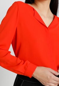 Blouse rouge à manches longues en tissu lisse avec un décolleté en V et un design subtil aux épaules, mettant en valeur la silhouette générale.