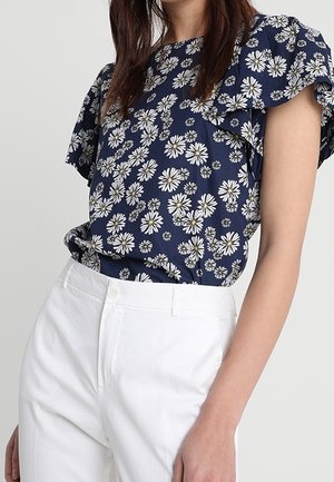 Vrouw draagt een navyblauwe blouse met witte madeliefjesprints en geplooide mouwen, gecombineerd met witte broeken met hoge taille.