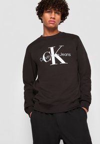 Svart hettegenser har et stort hvitt logo med teksten "Calvin Klein Jeans". Mykt stoff, rund hals og ribbestrikkede mansjetter, kombinert med svarte bukser.