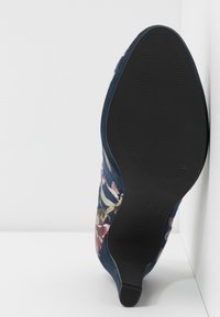 Semelle usée d'une chaussure à talons hauts à motif floral, avec un fond texturé noir et une taille 37, présentée sur un fond blanc.