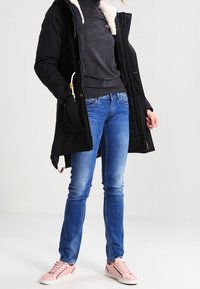 Svart parka-jacka med huva klädd i fuskpäls, grå tröja, blå jeans och ljusrosa sneakers. Avslappnad outfit med kontrasterande färger.