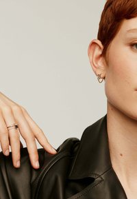 Pendiente de aro dorado con un diseño floral, que presenta una pequeña gema. Un dedo con un anillo de plata y oro descansa sobre una chaqueta de cuero negra.