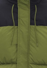 Puffjakke med olivengrønne og sorte paneler, der har to snore og en glat, quiltet tekstur. Lynlåsdetalje synlig.