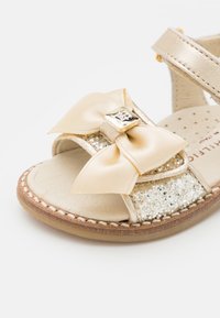 Sandalo in pelle dorata con un ampio fiocco in satin e accenti glitterati sulla fascia. La suola presenta un dettaglio cucito e una soletta testurizzata.