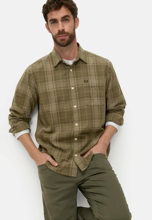 Mann mit Bart, der ein olivgrünes kariertes Hemd mit Knöpfen und passende grüne Hose trägt, die Hände in den Taschen, steht vor weißem Hintergrund.