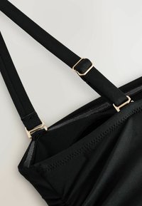 Reguliuojama juodos audinio liemenėlės petnešėlė su auksinės spalvos metaliniais slankikliais, pritvirtinta prie juodos liemenėlės kaušelio su siūtomis kraštinėmis.