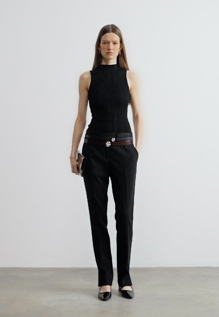 Femme debout contre un mur blanc, portant un haut noir sans manches, un pantalon noir avec deux ceintures, des chaussures noires à bouts pointus, tenant une petite pochette.