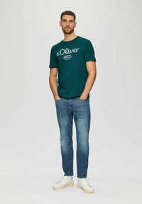 Türkise Baumwoll-T-Shirt mit weißem "s.Oliver"-Logo, kombiniert mit hellblauen Jeans und weißen Sneakers, vor einem neutralen Hintergrund stehend.