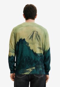 Stickad tröja i grönt och blått med ett mönster av berg och skog, med texturerat tyg och ribbade ärmslut och nederkant.