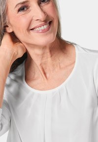 Weiße Bluse mit rundem Ausschnitt, weicher Textur und subtilen Faltendetails. Das Model hat lange graue Haare, eine entspannte Pose und lächelt.