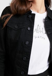 Žena v černé denimové bundě přes bílé tričko s černou liniovou kresbou panoramatu New Yorku a textem "York City."