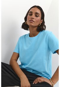 Lichtblauwe katoenen t-shirt met korte mouwen en een ronde hals, gecombineerd met zwarte broek, tegen een effen achtergrond.