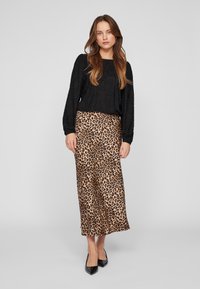 Nő áll egy fekete textúrájú, hosszú ujjú blúzban, leopárdmintás midi szoknyában és fekete, hegyes orrú magassarkúban egy sima szürke háttér előtt.