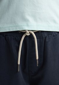 Un t-shirt en coton vert clair est rentré dans un pantalon bleu marine, doté d'un cordon de serrage avec une finition texturée et des embouts métalliques.