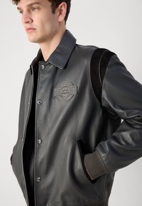 Zwarte leren varsityjack met zilveren knopen, geribbelde boorden en een getextureerde patch op de borst. Bevat zwart suède accenten op de schouders.