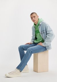 Νεαρός άνδρας φορώντας ανοιχτό μπλε τζιν μπουφάν, πράσινη κουκούλα, μπλε τζιν και λευκά αθλητικά παπούτσια ψηλής έκδοσης κάθεται πάνω σε ένα ξύλινο κουτί μπροστά σε λευκό φόντο.