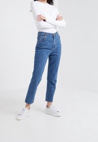 Jeans in denim blu a vita alta con gamba dritta, dotati di tasche frontali, cuciture sottili e chiusura con bottone. Indossati con sneakers bianche.