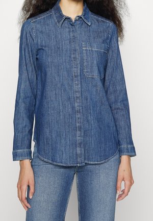 Žena v modré džínové košili s dlouhými rukávy a přední kapsou a modrých džínách, stojící před jednobarevným pozadím.