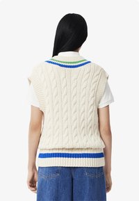 Maglione senza maniche a grosso filo di colore crema con colletto a righe verde e blu e una riga blu orizzontale all'orlo.