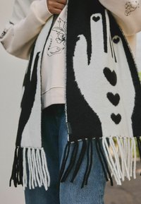 Écharpe tricotée en noir et blanc avec des motifs de mains et de cœurs, dotée de bords frangés. Le matériau semble doux et texturé, complétant les tenues décontractées.