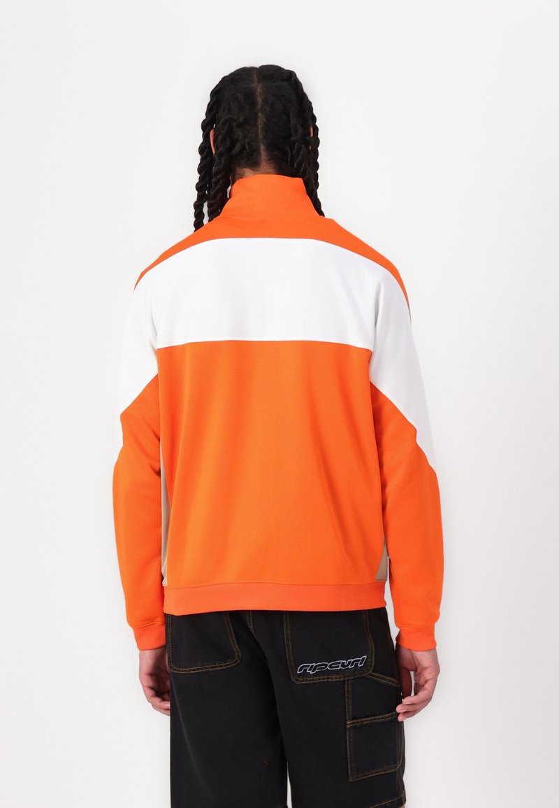 Orangene und weiße Zip-Jacke mit hohem Kragen. Glattes Material, Farbblock-Design, gerippte Bündchen und Saum. Getragen mit dunklen Cargo-Hosen.