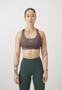 Soutien-gorge de sport marron à encolure ronde, avec un logo blanc, associé à des leggings taille haute vert foncé. Tissu lisse, design ajusté.