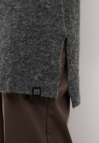 Grauer Wollpullover mit strukturierter Oberfläche, ausgestattet mit seitlichen Schlitzen und einem schwarzen Logotag am Saum, kombiniert mit braunen Hosen.