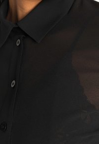 Chemise noire à col, avec des boutons et une texture transparente. Le tissu semble léger et lisse.