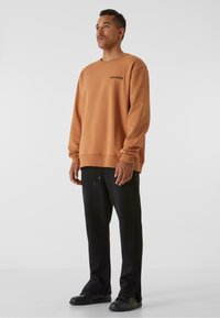 Felpa arancione oversize in cotone, con maniche lunghe e scollatura a girocollo, con la scritta "SWIM SENSE." stampata sul petto. Pantaloni neri.