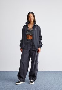 Chaqueta deportiva negra con rayas blancas, top corto de malla, pantalones de mezclilla anchos negros con bolsillos y zapatillas negras con suelas blancas.