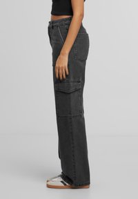 Personne portant un pantalon cargo noir taille haute et des baskets blanches avec des détails noirs, debout de côté devant un fond gris uni.