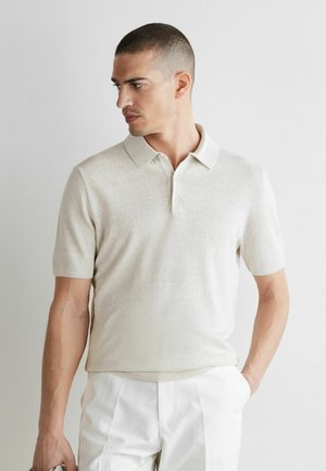 Mężczyzna w jasnobeżowej koszulce polo z krótkim rękawem i białych spodniach, patrzący w lewo z jedną ręką w kieszeni.