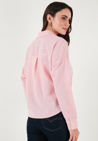 Light pink and white striped button-up shirt with long sleeves, collar, and back pleat detail. Soft fabric and curved hem design.
