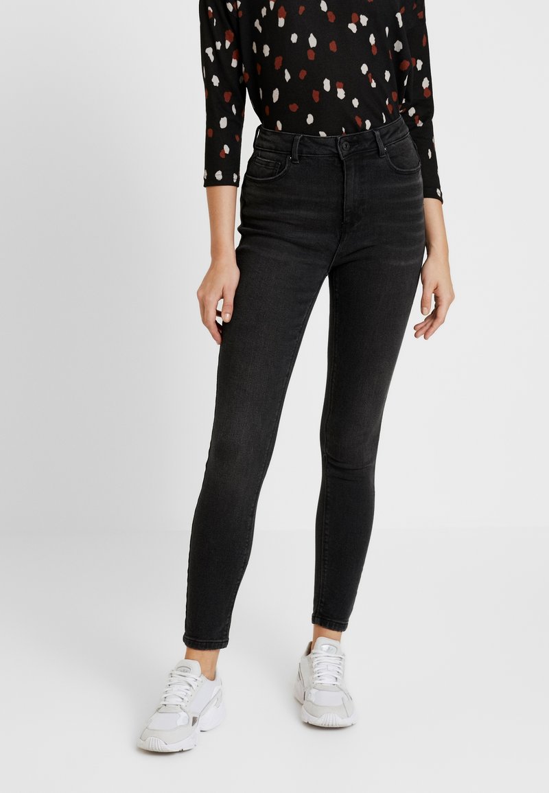 Svarta denim skinny jeans med hög midja, femficka design och subtila blekningar. Kombinerat med en mönstrad svart skjorta och vita sneakers.