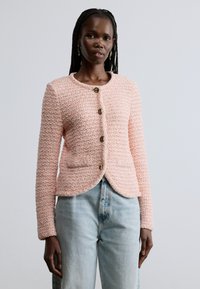 Világos rózsaszín texturált dzseki kerek nyakkal, öt arany tónusú gombbal és elülső zsebekkel, világoskék farmernadrág felett viselve.