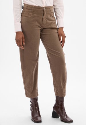 Bruine corduroy broek met een relaxte fit, voorzien van twee voorzakken en een knoopsluiting. Draagt met glanzende bruine enkellaarzen.