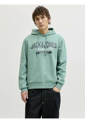 Ung mand iført en lys turkis hættetrøje med teksten "JACK & JONES" og mørke jeans, stående foran en ensfarvet hvid baggrund.