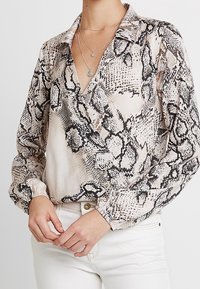 Donna che indossa una camicetta beige con stampa a serpente a maniche lunghe e scollatura a V, abbinata a collane di argento a strati e pantaloni bianchi a vita alta.
