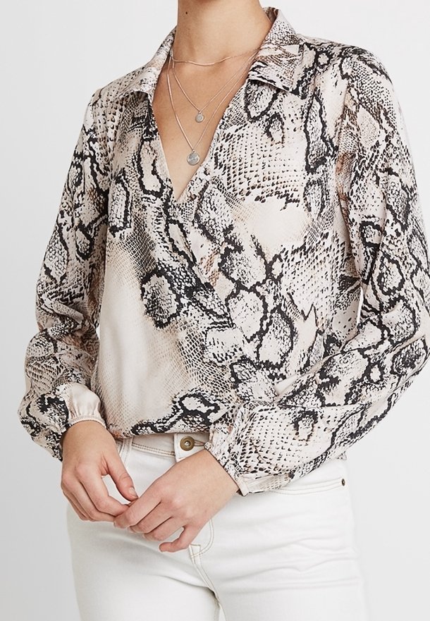 Donna che indossa una camicetta beige con stampa a serpente a maniche lunghe e scollatura a V, abbinata a collane di argento a strati e pantaloni bianchi a vita alta.
