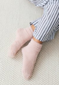 Pluizige, lichtroze sokken van zacht materiaal, rustend op een gestructureerd, lichtkleurig tapijt, naast grijs-wit gestreepte pyjamabroek.