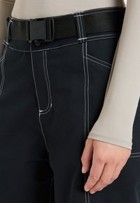 Pantalones negros con costuras blancas y un cinturón con una hebilla de plástico negra. La tela parece duradera y tiene una textura suave.