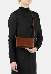 Une femme portant un haut à manches longues noir et une robe sans manches tient un sac en cuir marron à bandoulière avec une sangle d'épaule et un logo doré.