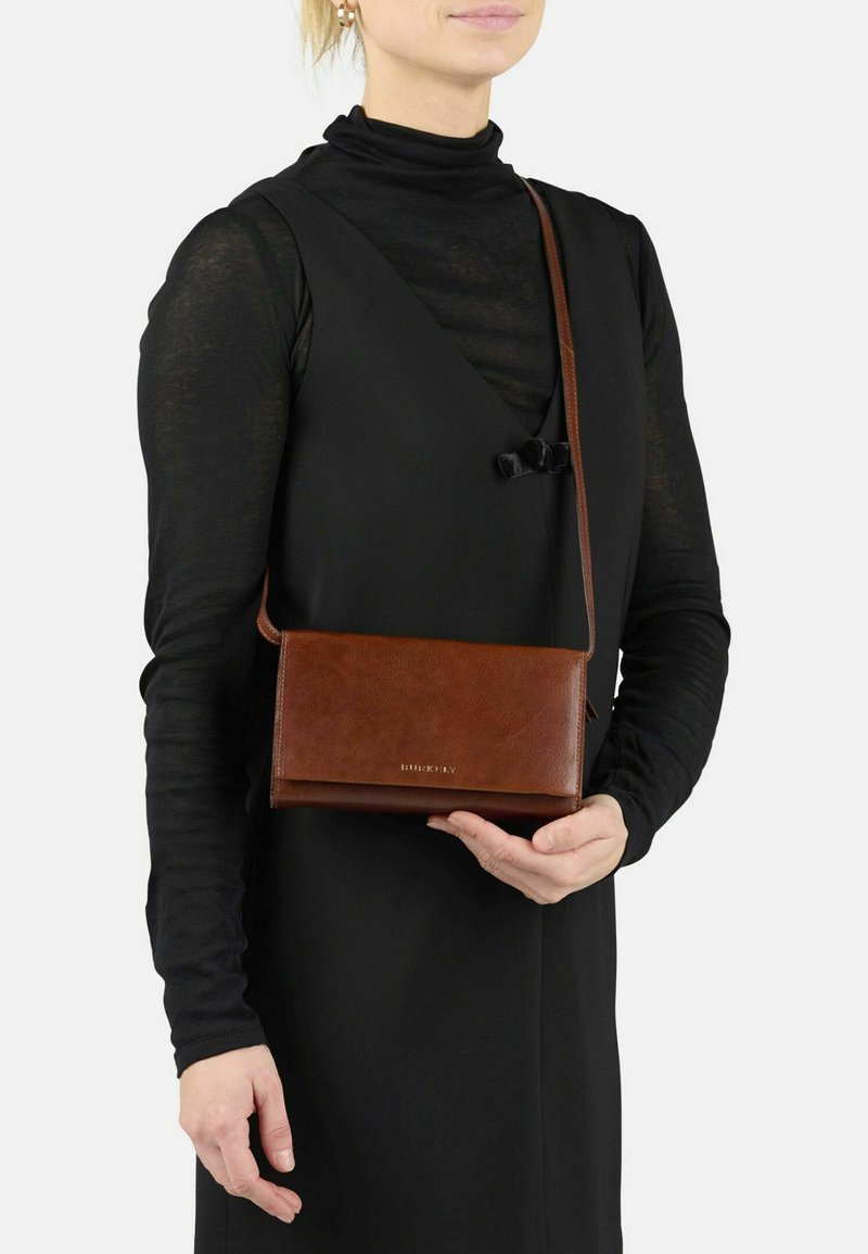 Une femme portant un haut à manches longues noir et une robe sans manches tient un sac en cuir marron à bandoulière avec une sangle d'épaule et un logo doré.
