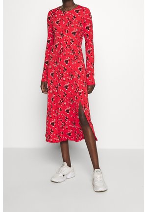 Vrouw draagt een rode midi-jurk met lange mouwen en bloemenprint, met een zijsplit, en witte sneakers, staand tegen een effen witte achtergrond.